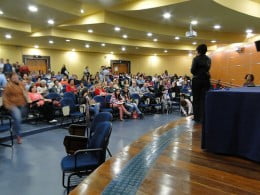 A imagem mostra um palestrante conversando com um auditório cheio.