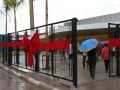 Foto mostra portões com um laço vermelho e pessoas entrando com guarda-chuvas