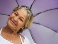 Foto mostra senhora sorrindo segurando um guarda-chuva lilás