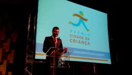 Pedro Hartung falando ao microfone atrás de um púlpito transparente. Atrás, um telão com o texto: Prêmio Cidade da Criança