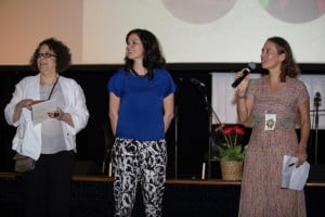 Foto mostra três mulheres e uma falando ao microfone