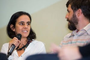 Foto mostra Renata Meirelles falando ao microfone enquanto olha para David Reeks