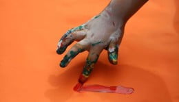 Mão fazendo um risco vermelho de tinta, em cima de um papel laranja.