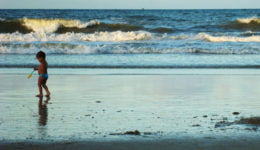 Imagem de uma praia com uma criança parada ao lado esquerdo.
