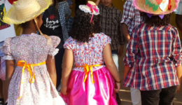 Na imagem, três crianças aparecem de costas e mãos dadas, vestindo roupas de festa de junina.