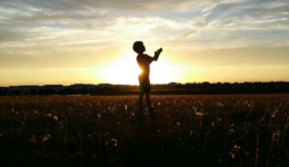 Na foto, a sombra de um menino, em um campo, com as mãos voltadas ao céu.