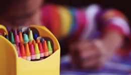 Uma caixa cheia de gizes colorido, e ao fundo uma criança pintando.
