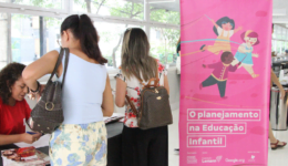 Foto de banner com o texto "O Planejamento da Educação Infantil". Ao lado, duas mulheres pegando suas credenciais em uma mesa