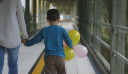 Imagem do documentário miradas. Na imagem, um adulto e um menino caminham com bexigas amarelas e rosas na mão. Os dois estão de costas andando por um corredor.