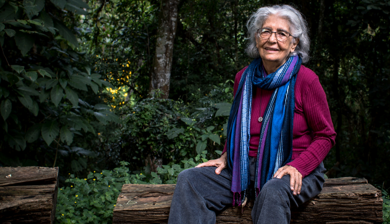 Foto mostra Lydia Hortélio sentada em um banco no meio da natureza