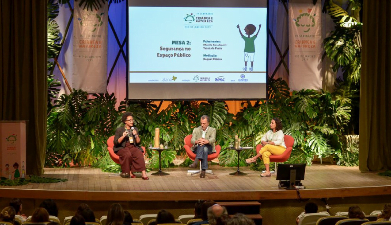 Foto do seminário "Infâncias e Naturezas" mostra palco com um telão e três palestrantes sentados de frente para a plateia