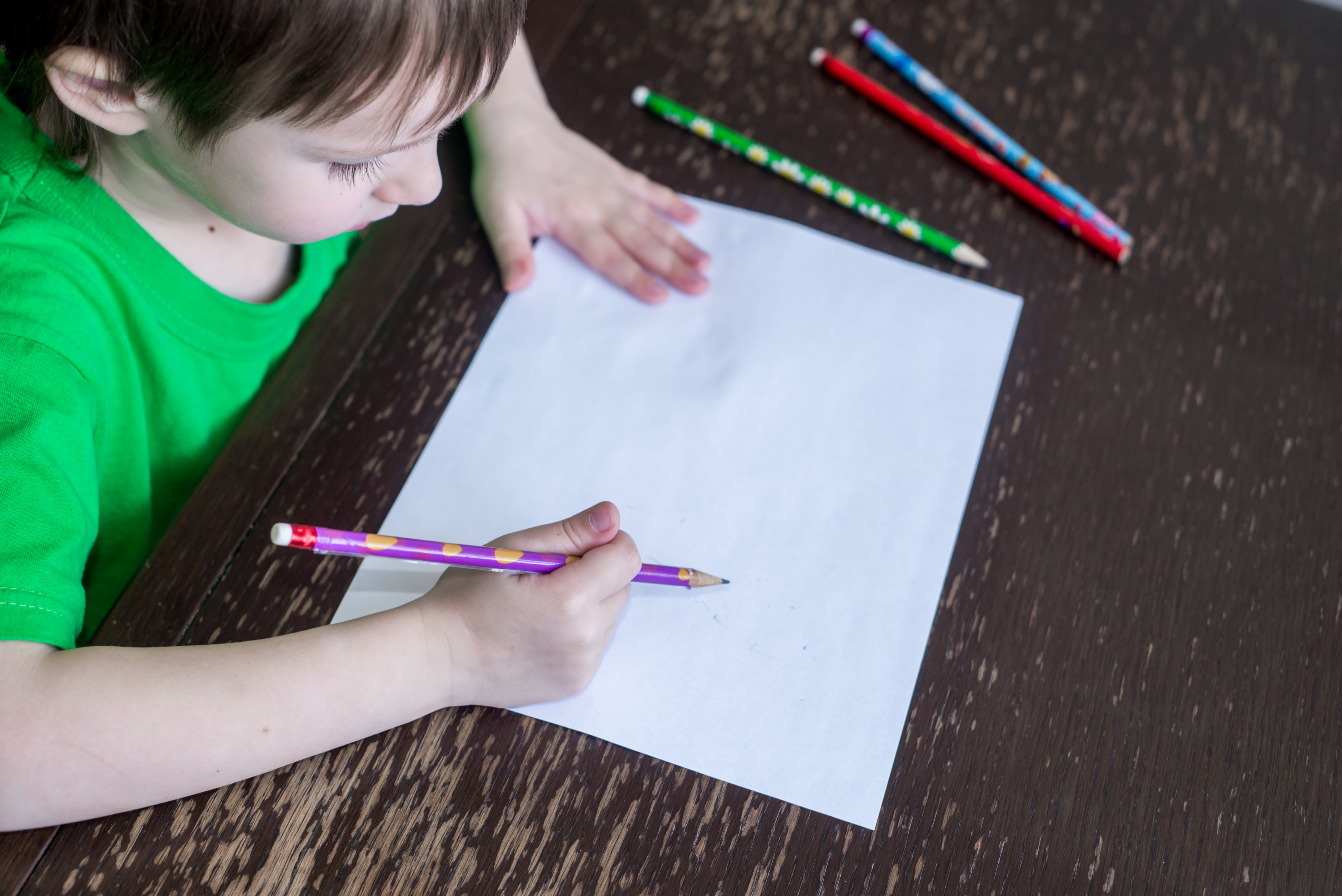 Foto de um menino branco em ensino domiciliar. Ele segura um lápis, diante de uma folha sulfite que está apoiada em uma mesa.