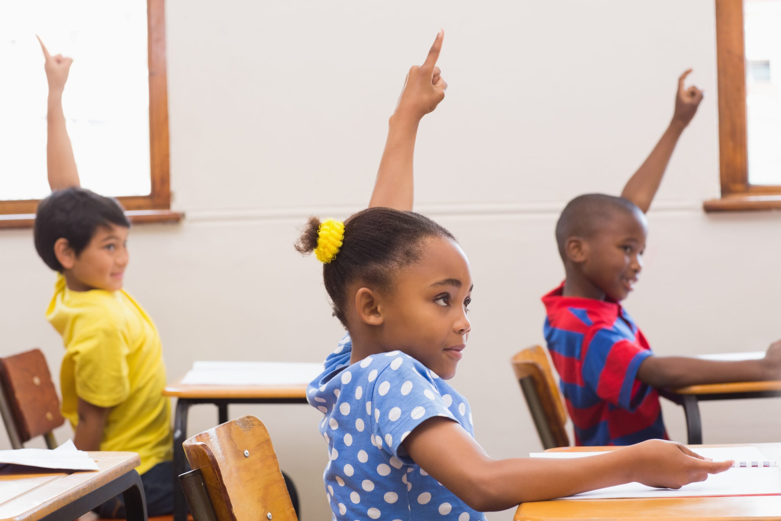 Foto mostra várias crianças em sala de aula com a mão levantada