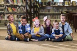 Foto mostra várias crianças sorrindo sentadas no chão da biblioteca de uma escola
