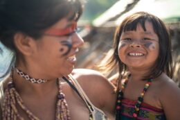 Foto mostra mulher adulta segurando criança no colo. Ambas são indígenas e estão sorrindo. Literatura indígena.