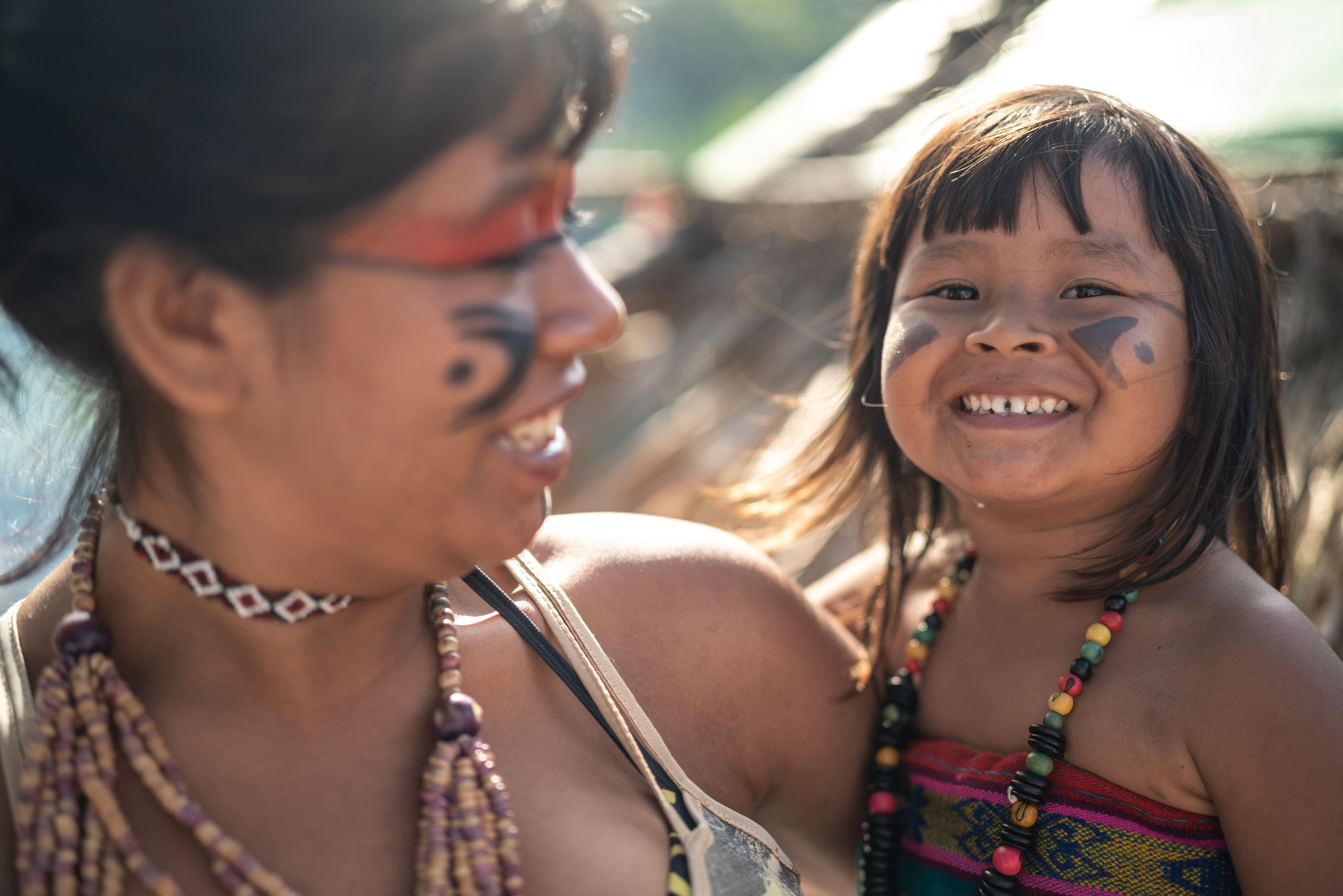 Foto mostra mulher adulta segurando criança no colo. Ambas são indígenas e estão sorrindo