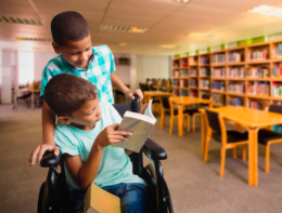 Foto mostra duas crianças lendo em biblioteca, uma delas está em uma cadeira de rodas. Imagem representa a educação inclusiva