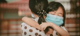 Na foto, uma menina branca de máscara de proteção abraça emocionada uma mulher adulta, sugerindo os efeitos dos impactos da gestão da pandemia sobre crianças e adolescentes.