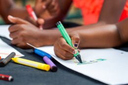 Ensino domiciliar: foto mostra o detalhe das mãos de duas crianças negras desenhando no caderno em uma mesa.