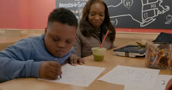 Alana Down Syndrome Center: na imagem, uma criança negra com síndrome de Down desenha em uma folha sulfite. Ao fundo, uma mulher negra o observa.