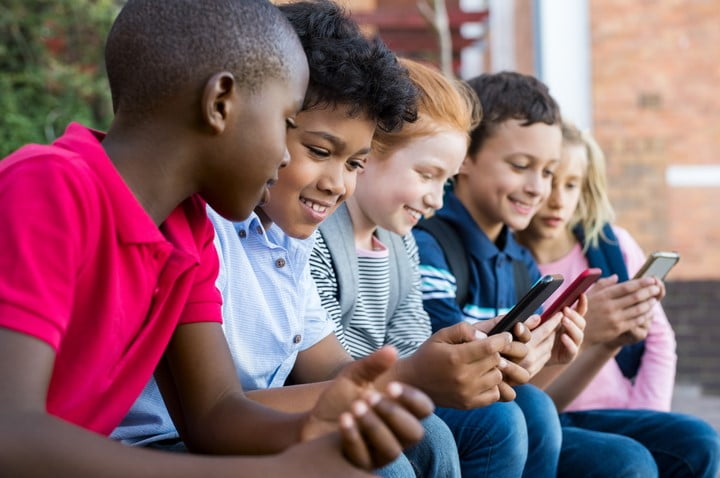 Na imagem, crianças brancas e negras estão sentadas lado a lado, olhando e mexendo no celular.