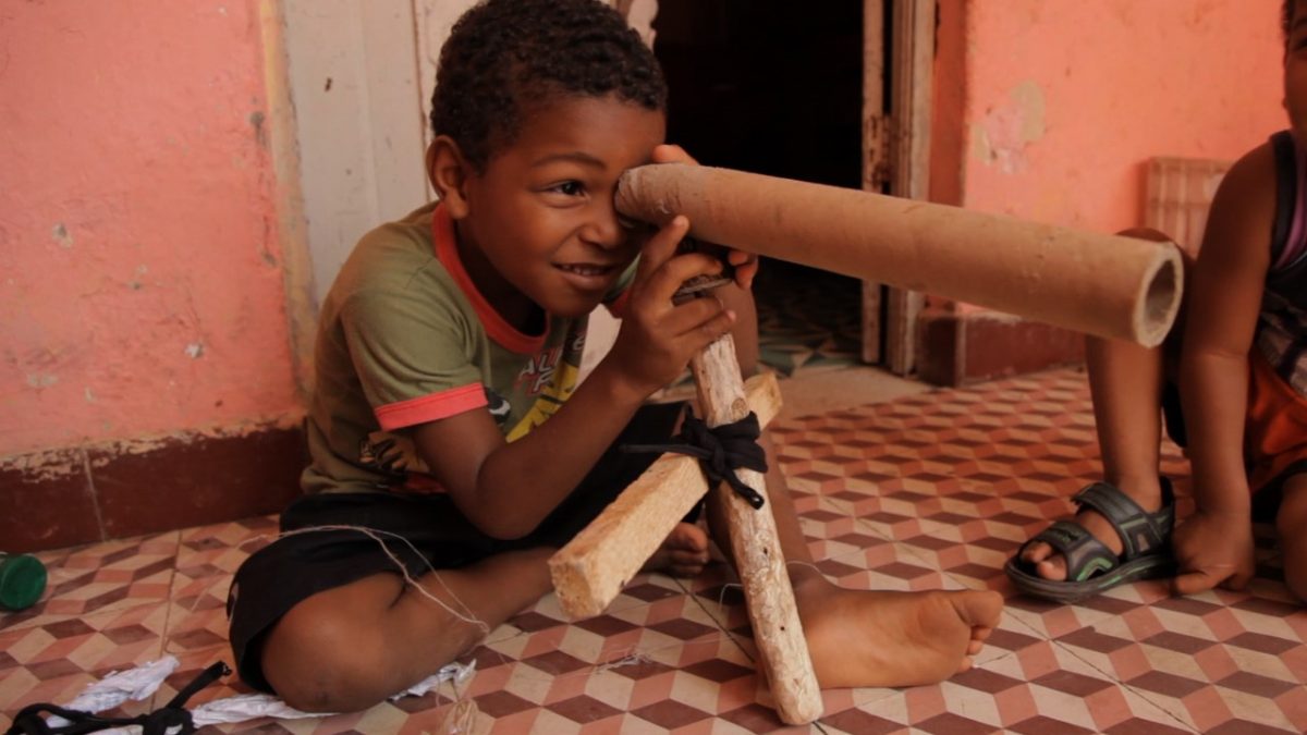 Na imagem, uma criança negra olha através de uma luneta feita de cilindro de papelão.