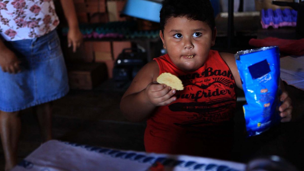 Muito Além do Peso: na imagem, uma criança de blusa vermelha come batata frita de um pacote de salgadinho.