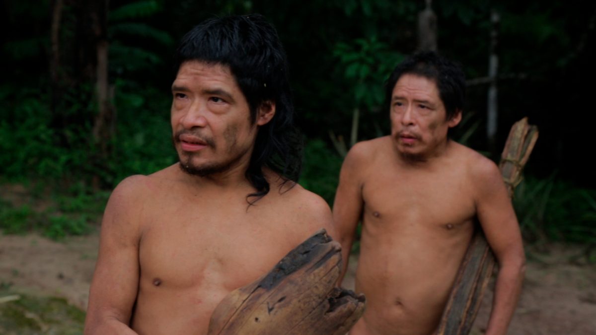 Cenas do documentário PIRIPKURA. Na imagem, dois homens indígenas caminham em uma floresta. Eles carregam nas mãos troncos de árvores.