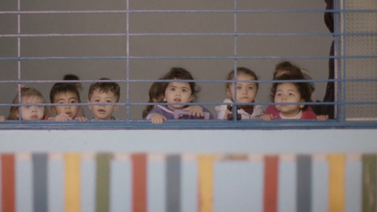 Cena de "O Começo da Vida 2: Lá fora". Na imagem, crianças observam a vista de fora através de uma janela com grades. Elas estão em uma sala de aula.