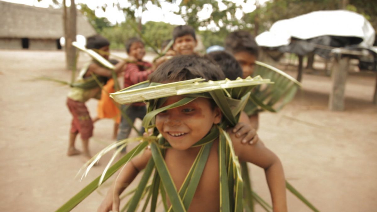 Cena do filme Terreiros do Brincar. Na imagem, crianças indígenas brincam de trenzinho. Elas vestem roupas feitas com faixas de folhas de bananeira.