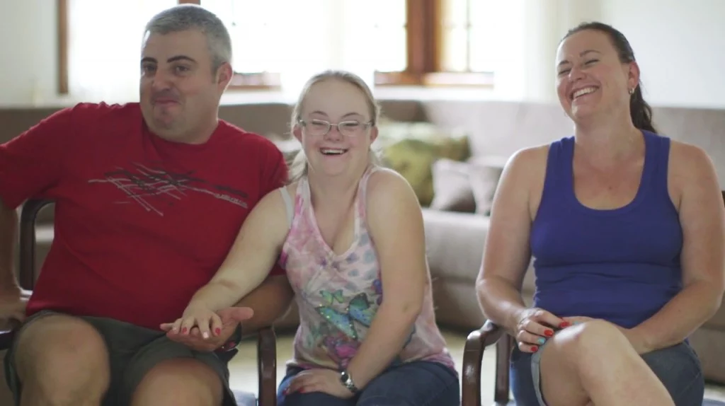 Cena do filme Outro Olhar – Uma nova perspectiva. Na imagem, uma adolescente com síndrome de Down está sentada em uma sala ao lado de dois adultos sem deficiência. Eles estão sentados em uma sala de estar e sorriem.
