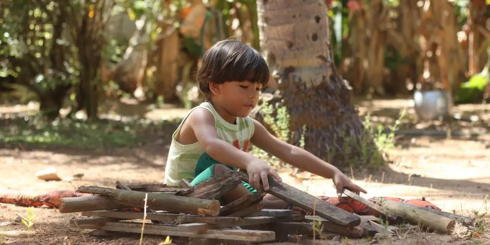 Criança e Natureza: na imagem, uma criança brinca com um montinho de pedaços de madeira e está sentada em um chão de terra.
