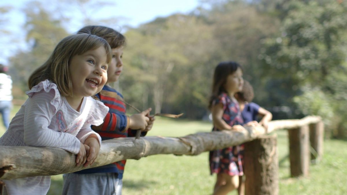 Cena do filme O Começo da Vida: na imagem, quatro crianças sorriem. Elas estão com o corpo inclinado sobre uma cerca de madeira.