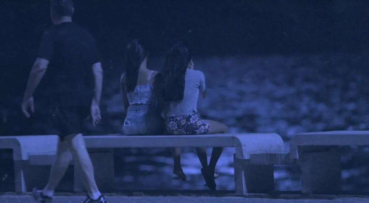 Na imagem, cena do documentário Um Crime Entre Nós. Duas adolescentes estão de costas, sentadas em um banco que fica de frente para o rio. Um homem passa ao fundo. Há um filtro azulado sobre a imagem.
