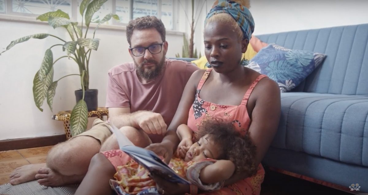Cena do documentário "Por Uma Educação Antirracista". Na imagem, uma mulher negra, um homem branco e uma criança negra leem um livro juntos, na sala de estar de uma casa.