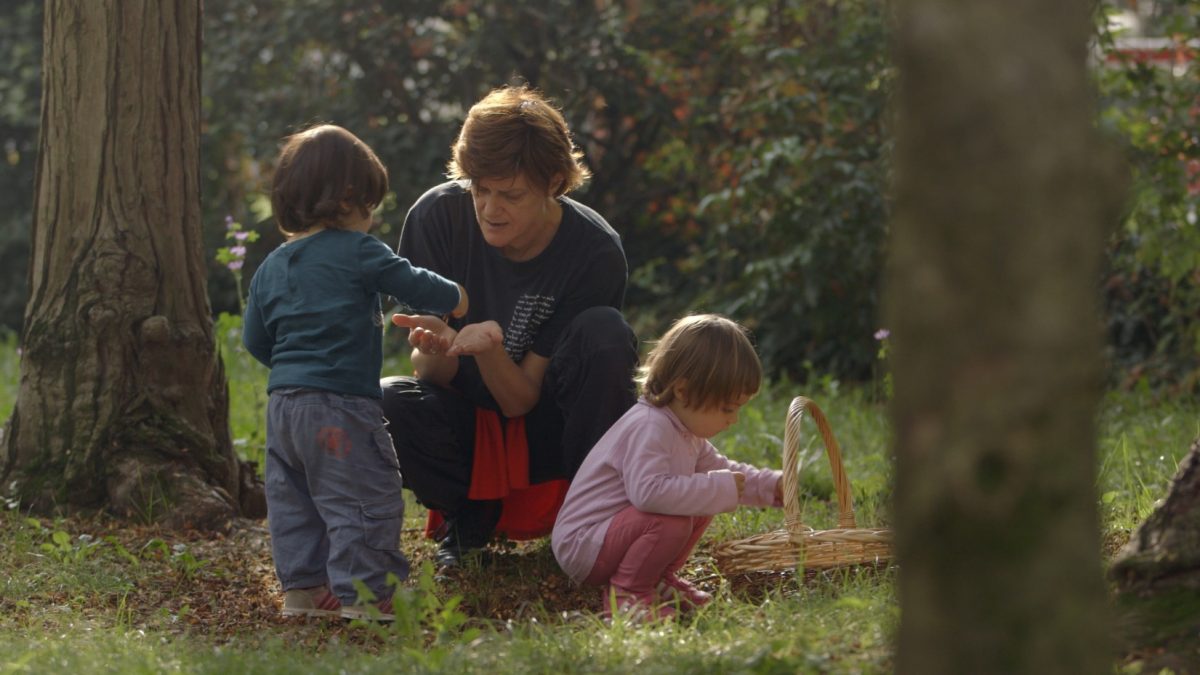 Cena de "O Começo da Vida – A Série". Na imagem, uma mulher branca colhe gravetos do chão com a ajuda de duas crianças pequenas. Na frente delas, há uma cesta de palha.