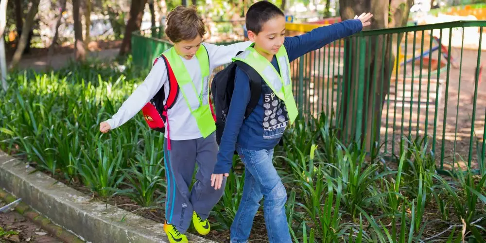 Caminhando Juntos até a Escola: na imagem, duas crianças que usam coletes de segurança caminham em fila sobre a sarjeta de uma praça.