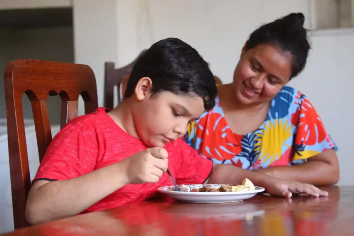 Obesidade em Crianças e Adolescentes: na imagem, uma criança se alimenta na mesa supervisionada por uma mulher sorridente.