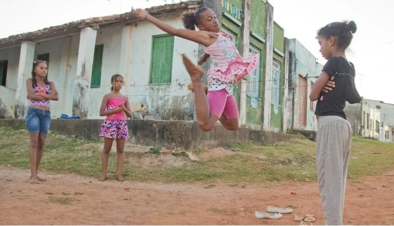 Na imagem, uma criança negra brinca com elástico em uma rua com chão de terra.