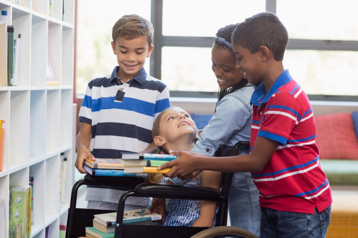 Pela Inclusão: em uma biblioteca, três crianças se reúnem em torno de uma criança com deficiência, que está ao centro da foto. Elas se olham e trocam sorrisos.