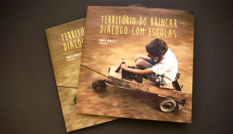 Dois exemplares do livro "Território do Brincar - Diálogo com Escolas". Na foto da capa, uma criança branca brinca em um carrinho de rolimã.