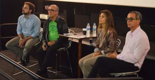 Realização de Cine Debates com educadores. Na imagem, quatro adultos brancos estão no palco em uma mesa de debate.