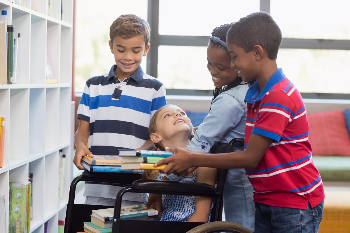 Na foto, quatro crianças brancas e negras interagem em uma biblioteca. Uma delas está em uma cadeira de rodas.