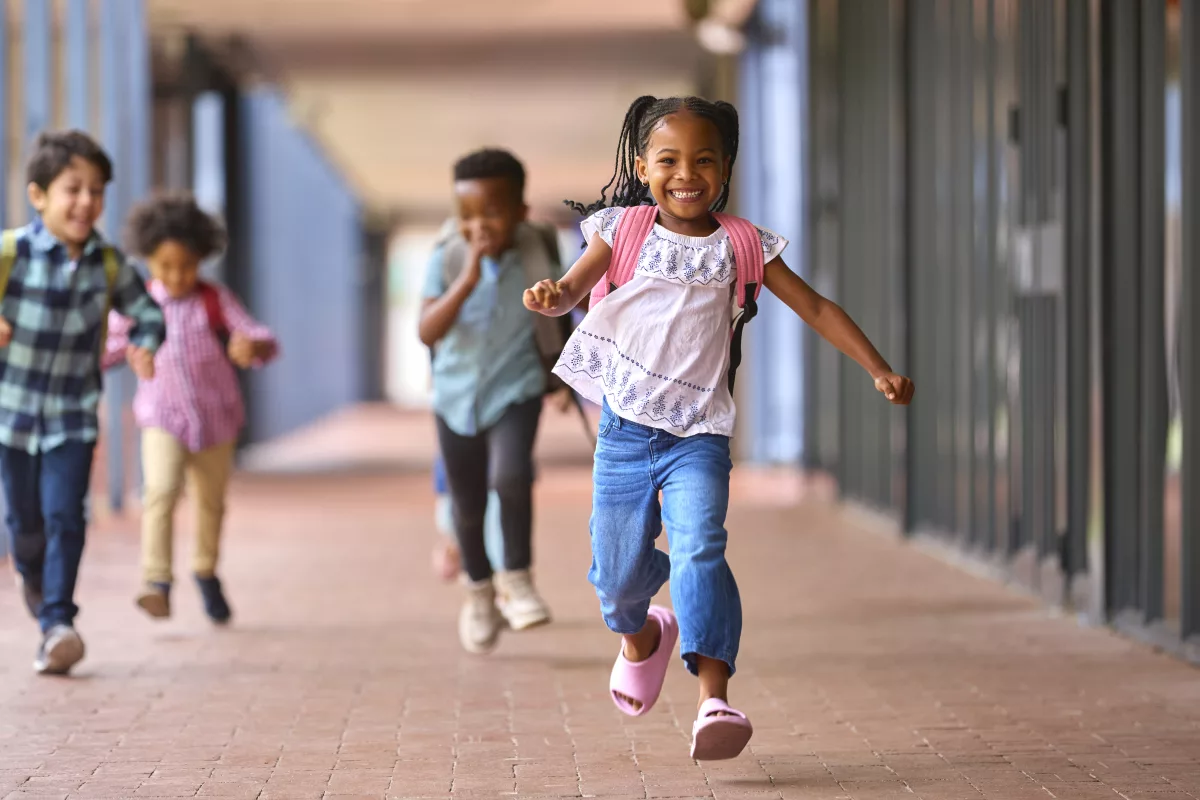 Lei 10.639: quatro crianças correm em um corredor escolar. Em destaque, uma menina negra, que está sorrindo e tem uma mochila rosa nas costas.
