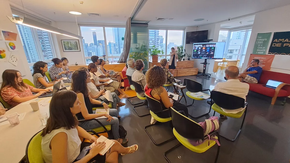 COP28: no escritório do Alana, grupo sentado em cadeiras assiste a uma palestra de uma pessoa que está em pé. Ao fundo, os prédios de São Paulo.