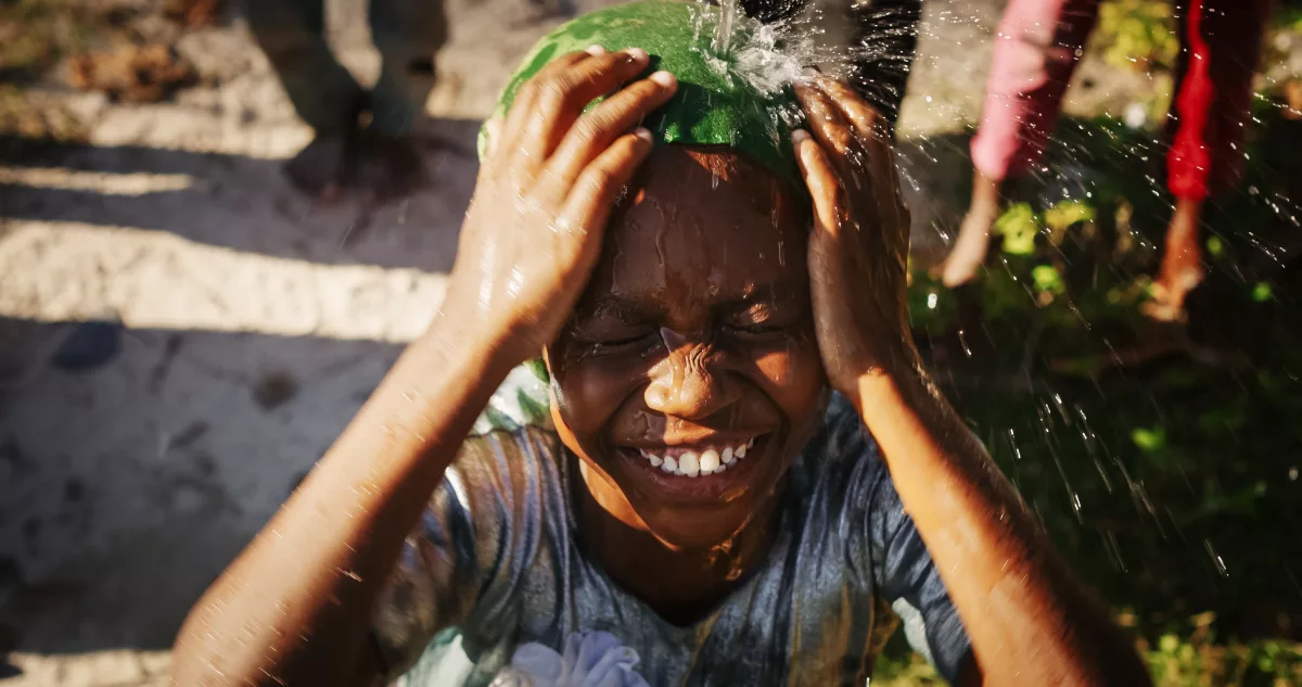 COP28: na imagem, uma criança negra toma uma ducha de em um espaço aberto e ensolorado. Ela sorri.