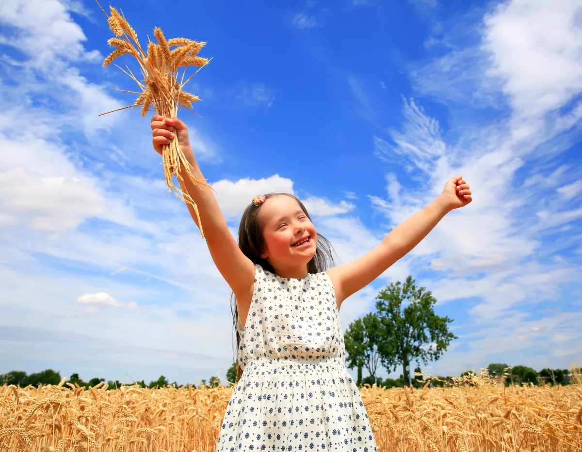 Na foto, uma criança branca com síndrome de Down abre os braços e sorri em um campo. Ela segura em uma das mãos um ramo de trigo.