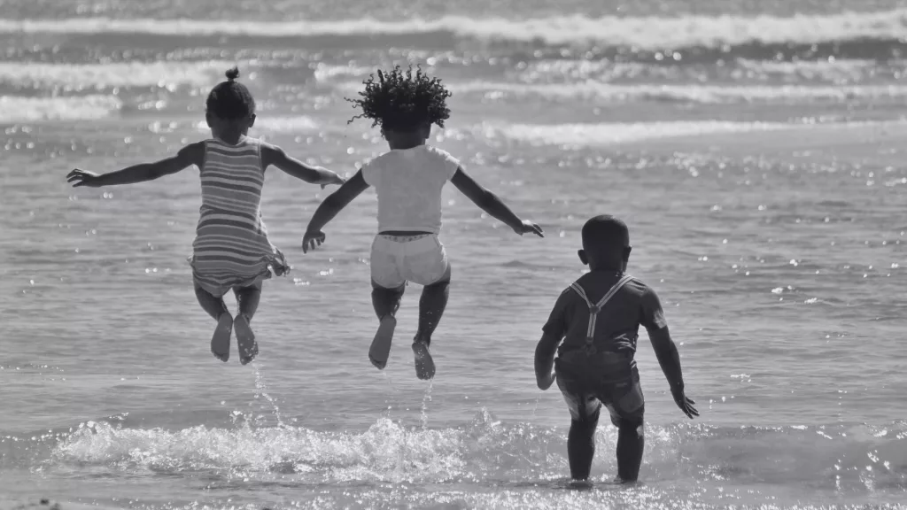 Foto de três crianças negras na praia. Elas estão de costas, pulando no mar. Imagem ilustra matéria "Instituto Alana participa de audiência no STF que volta a proibir apreensões ilegais de adolescentes", sobre a Operação Verão no Rio de Janeiro.