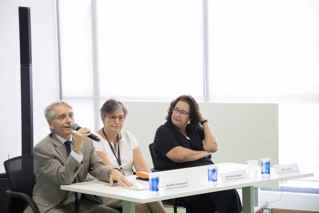 Na foto, Aluísio Segurado, Suzana Torresi e Ana Lanna. Os três são pessoas brancas e estão sentados em uma mesa. Na ponta esquerda, Aluísio Seguado está vestido com um terno cinza e fala em um microfone.