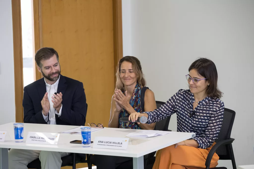 Na foto, Pedro Hartung, Isabella Henrique e Ana Lucia Villela. Os três são pessoas brancas e sorriem. Na ponta direita, Ana Lucia Villela, presidente do Alana, coloca o microfone em cima da mesa. 